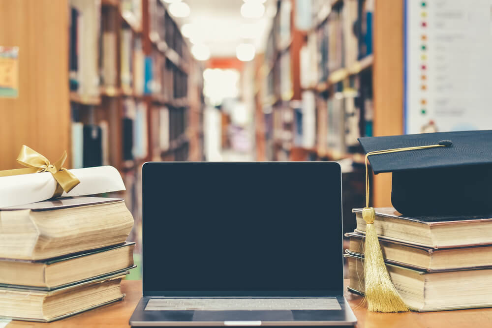 computer in biblioteca, sopra i libri c'è un tocco e una laurea per rappresentare differenze tra master e laurea magistrale