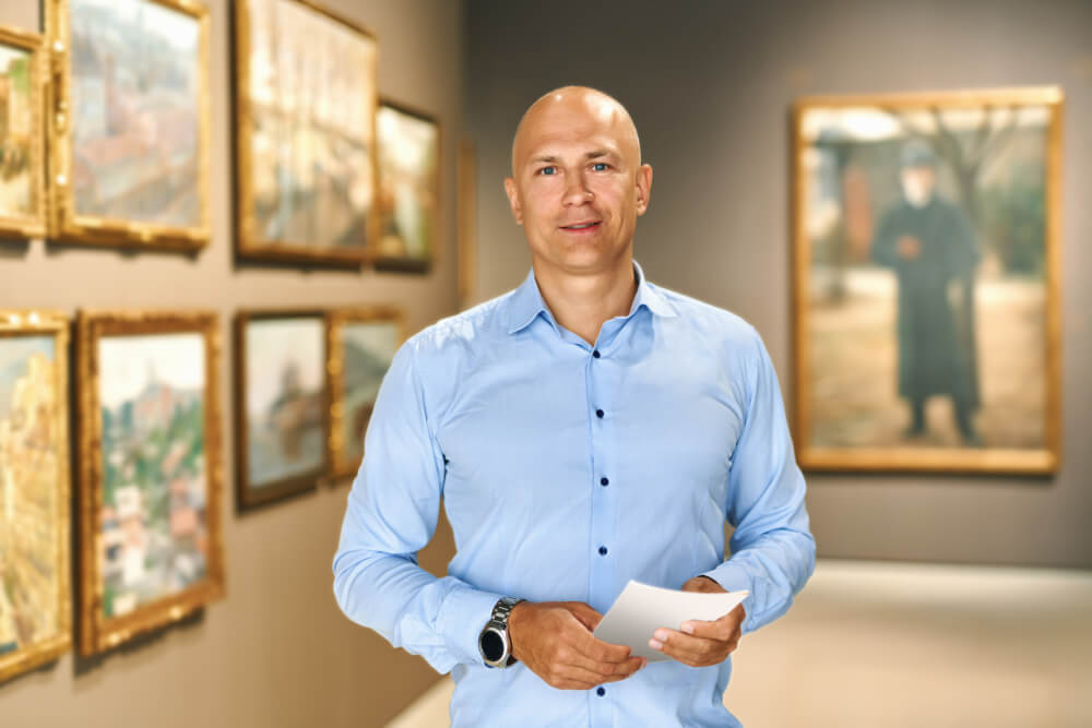 Portrait,Of,Man,With,A,Guide,Looking,At,Pictures,At Esperto di beni culturali in camicia celeste nella galleria di un museo con tanti quadri affissi alle pareti
