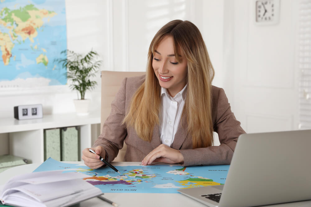 Happy,Manager,Working,At,Desk,In,Travel,Agency Manager del turismo osserva una cartina e indica un punto sulla mappa con il pc davanti