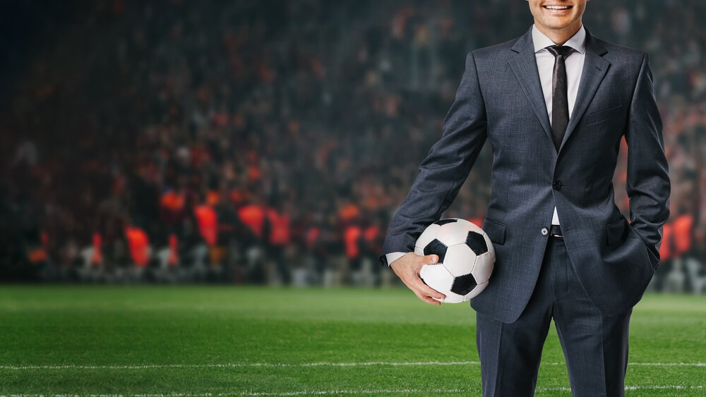 Smiling,Businessman,In,Formal,Wear,With,Soccer,Ball,In,The Football manager sul campo di calcio con completo nero e cravatta