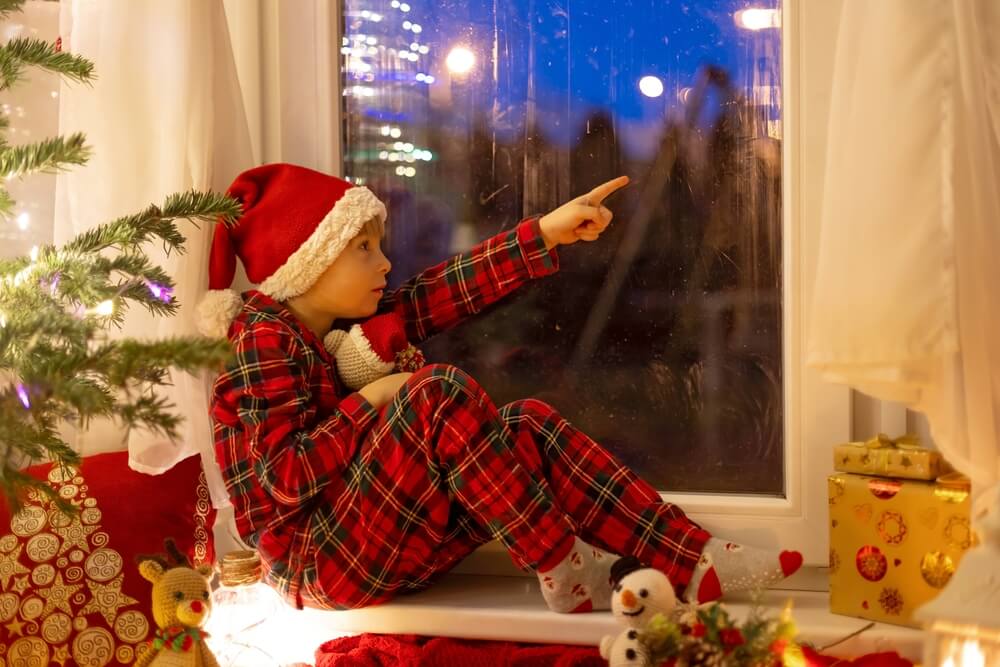Cute,Child,,Sitting,On,A,Window,,Looking,Outdoors,For,Santa Bambino con cappellino da babbo natale è seduto sul davanzale e indica dalla finestra