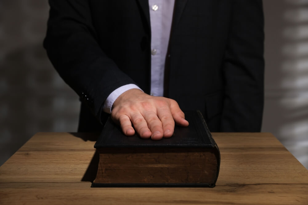 Man,Taking,Oath,With,His,Hand,On,Bible,At,Wooden Uomo che giura con la mano sulla Bibbia