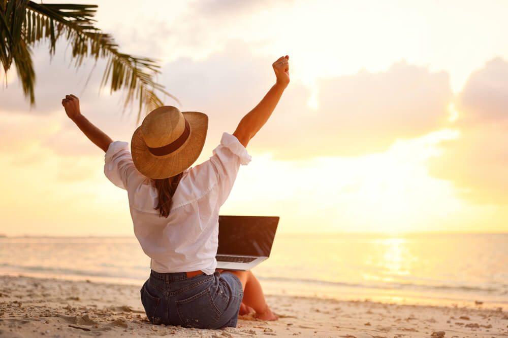 Ragazza di spalle, in spiaggia, con cappello di paglia a tesa larga regge laptop sulle ginocchia e si sgranchisce