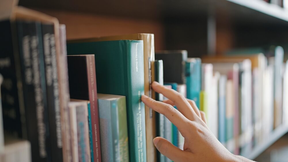 Close,Up,Hand,Search,For,Book,On,Bookshelves,In,The Primo piano di mano in libreria che scorre tra i volumi sugli scaffali