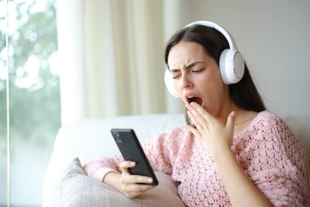 Bored,Woman,Wearing,Headphone,Yawning,Listening,Audio,On,Phone,At Donna ascolta la musica con cuffie alle orecchi e sbadiglia annoiata