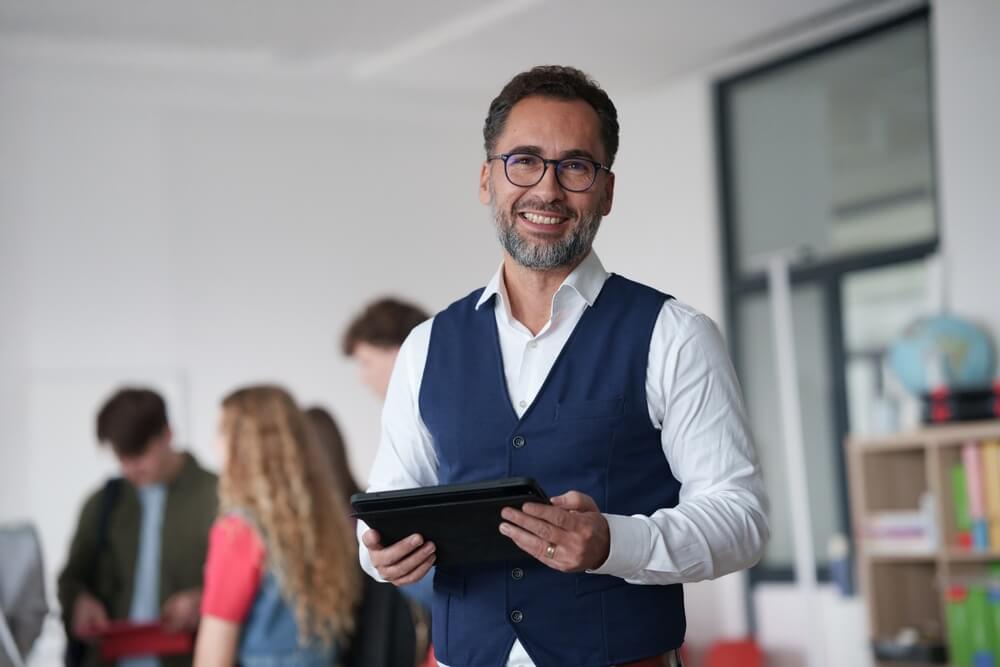 Teacher,Holding,A,Tablet,In,Modern,High,School,Classroom. Insegnante sorride mentre tiene in mano un tablet in una classe di liceo