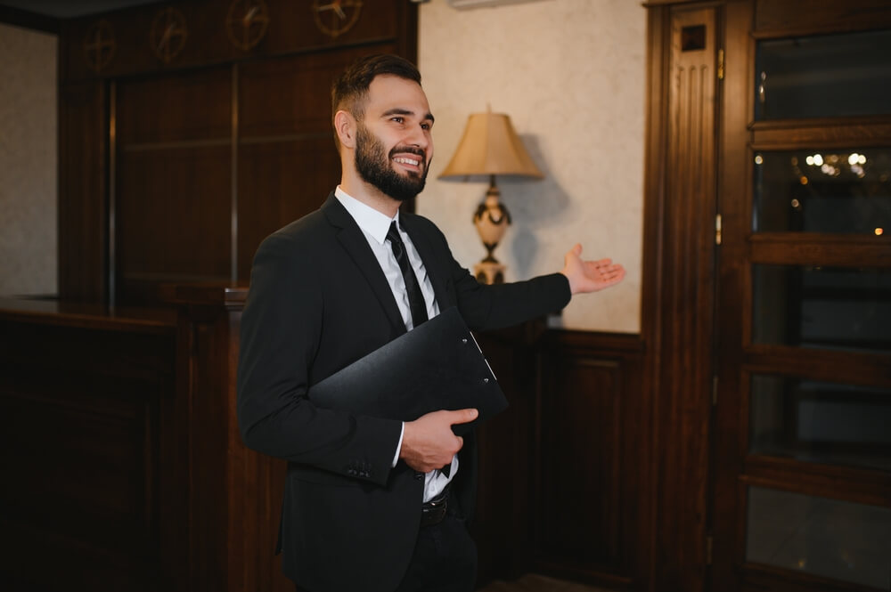 Smiling,Man,In,A,Suit,Working,As,A,Hotel,Receptionist, Uomo sorridente in albergo accoglie gli ospiti. Direttore o receptionist.
