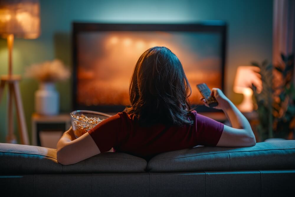 Donna di spalle sul divano di casa guarda la tv e mangia popcorn
