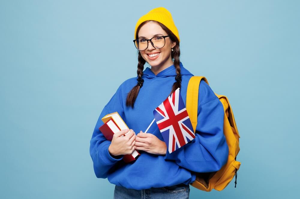 Giovane ragazza con zaino giallo in spalla che rappresenta il concetto di studente di lingue tiene la bandierina della Gran Bretagna