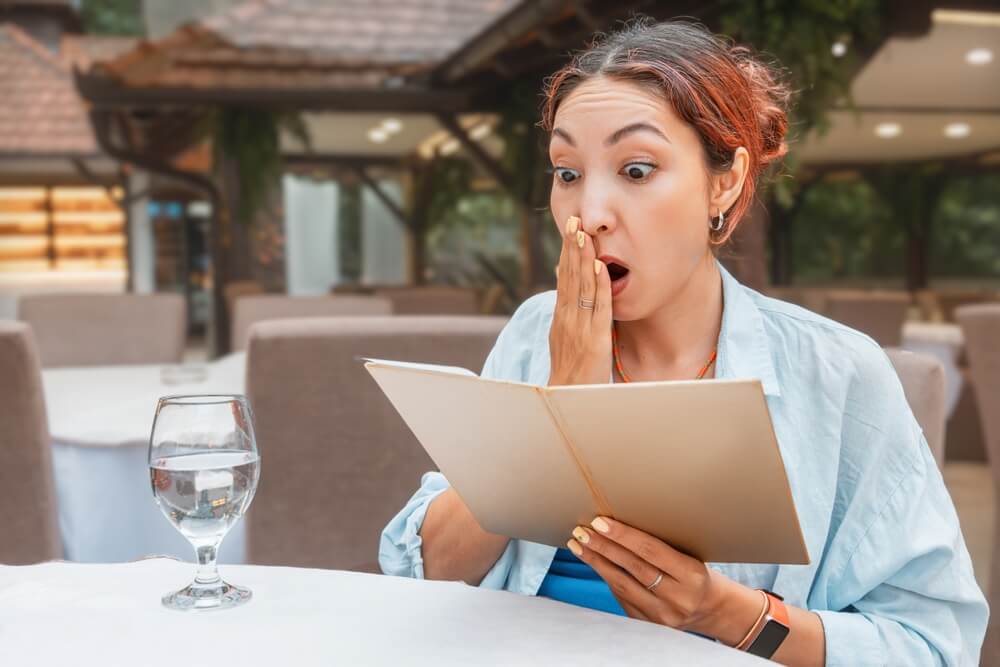 Woman,Looking,Surprised,And,Shocked,While,Holding,A,Menu,,Realizing Donna sbalordita legge il conto del ristorante